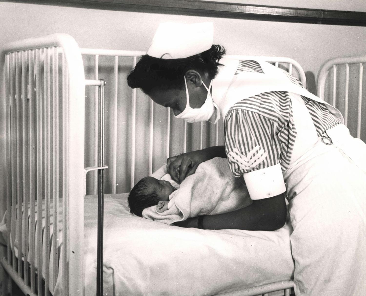A mid-twentieth-century photograph of a nurse tending a newborn in a hospital crib